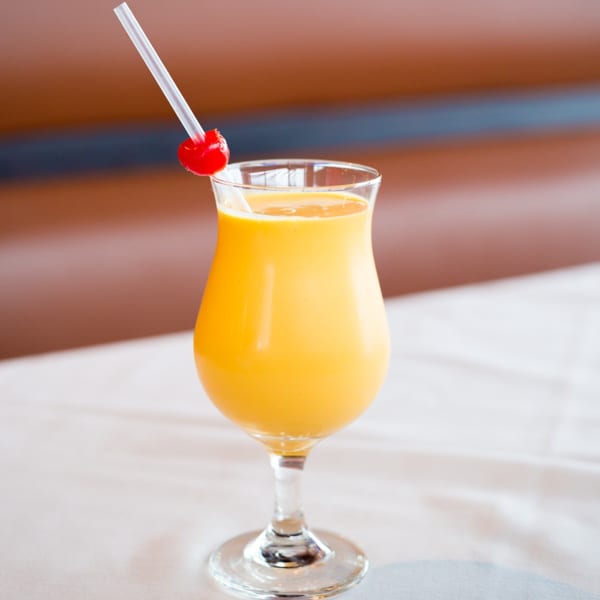A vibrant yellow mango lassi in a stemmed glass, garnished with a red cherry and straw.