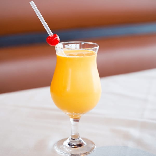 A vibrant yellow mango lassi in a hurricane glass, garnished with a red cherry and straw.