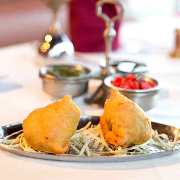 Two golden-fried samosas on a silver platter with shredded garnish, accompanied by two dipping sauces.