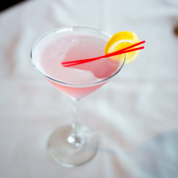 Pink cocktail in a martini glass with a lemon slice and two red straws.