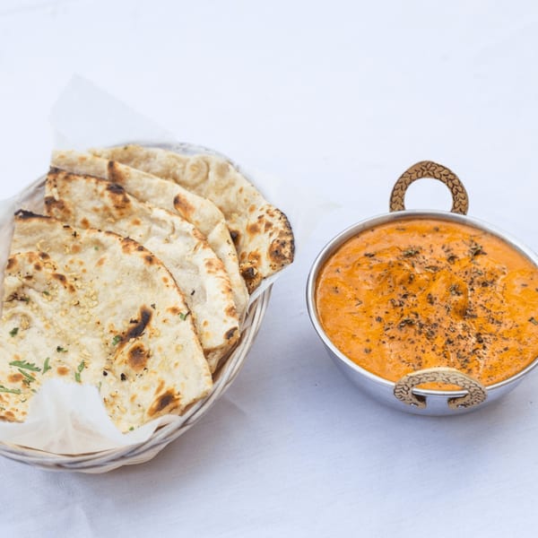 A basket of naan bread next to a silver bowl of orange Indian curry on a white tablecloth.